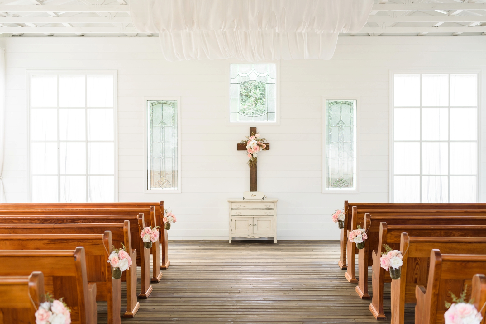 The wedding ceremony filled with white tulle and floral filled pews by Sarah and Ben Photography