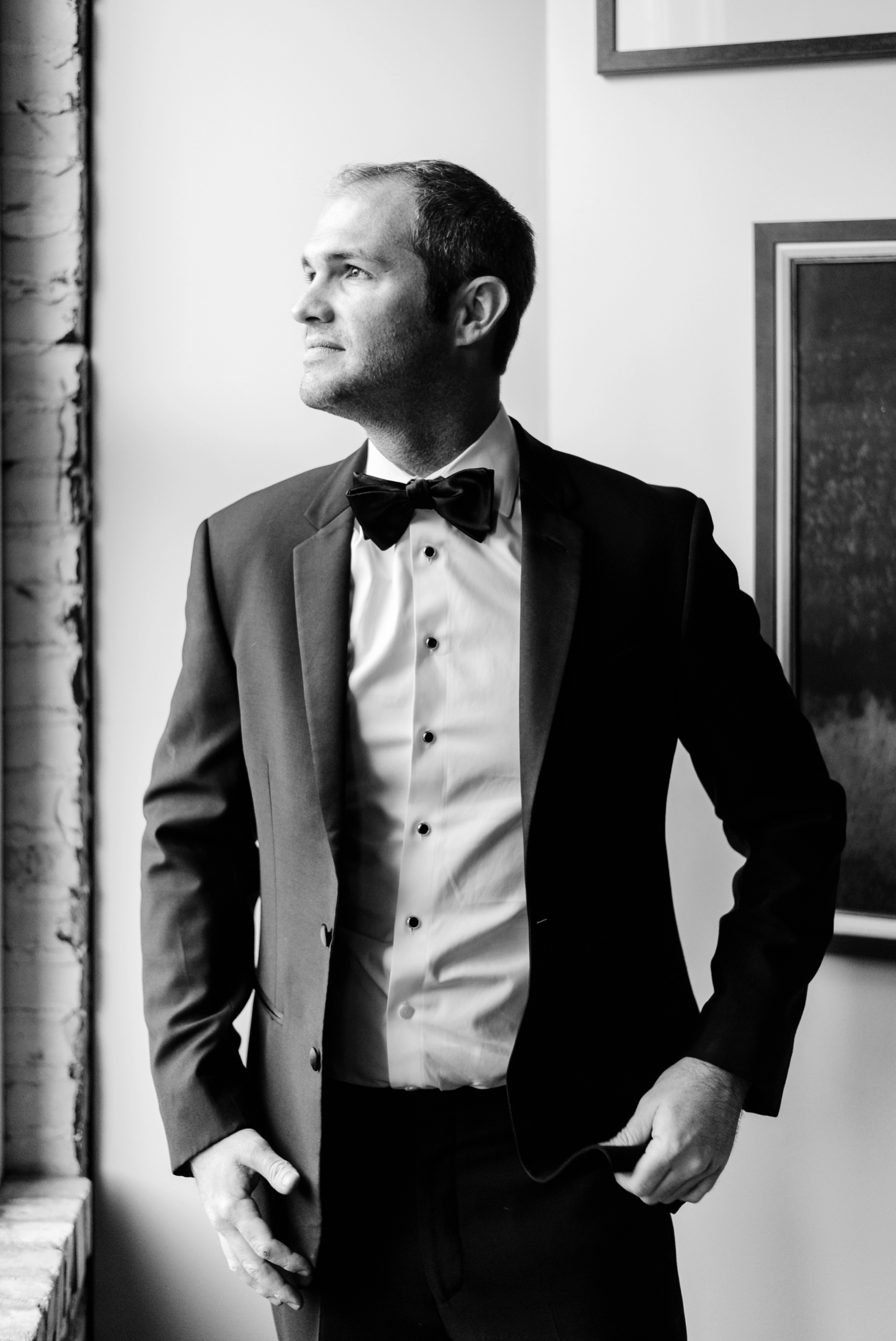 The Groom in a timeless black and white image looking out a window at the clouds by Sarah and Ben Photography
