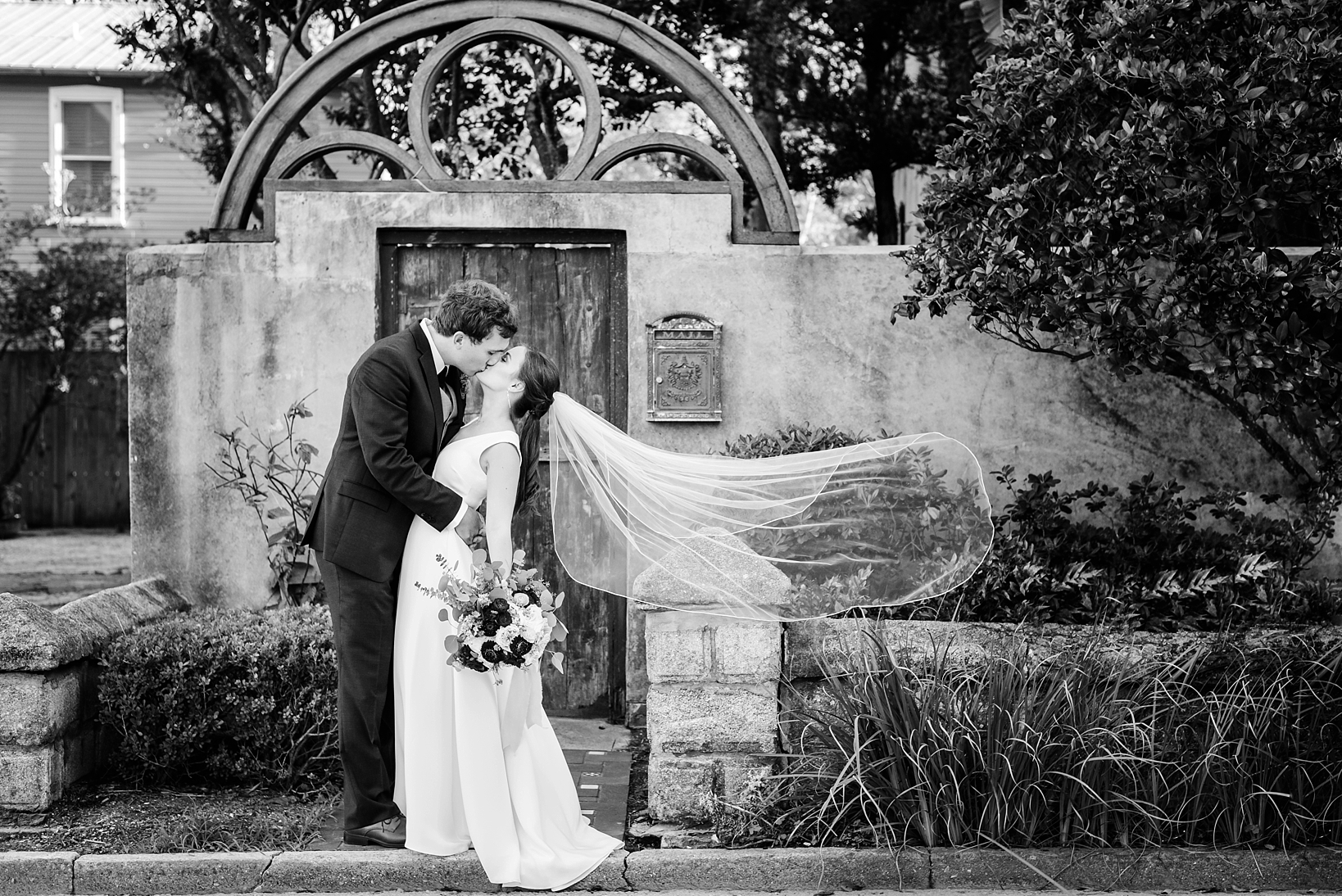 Black and white image of a bride and groom kissing while her veil blows in the wind