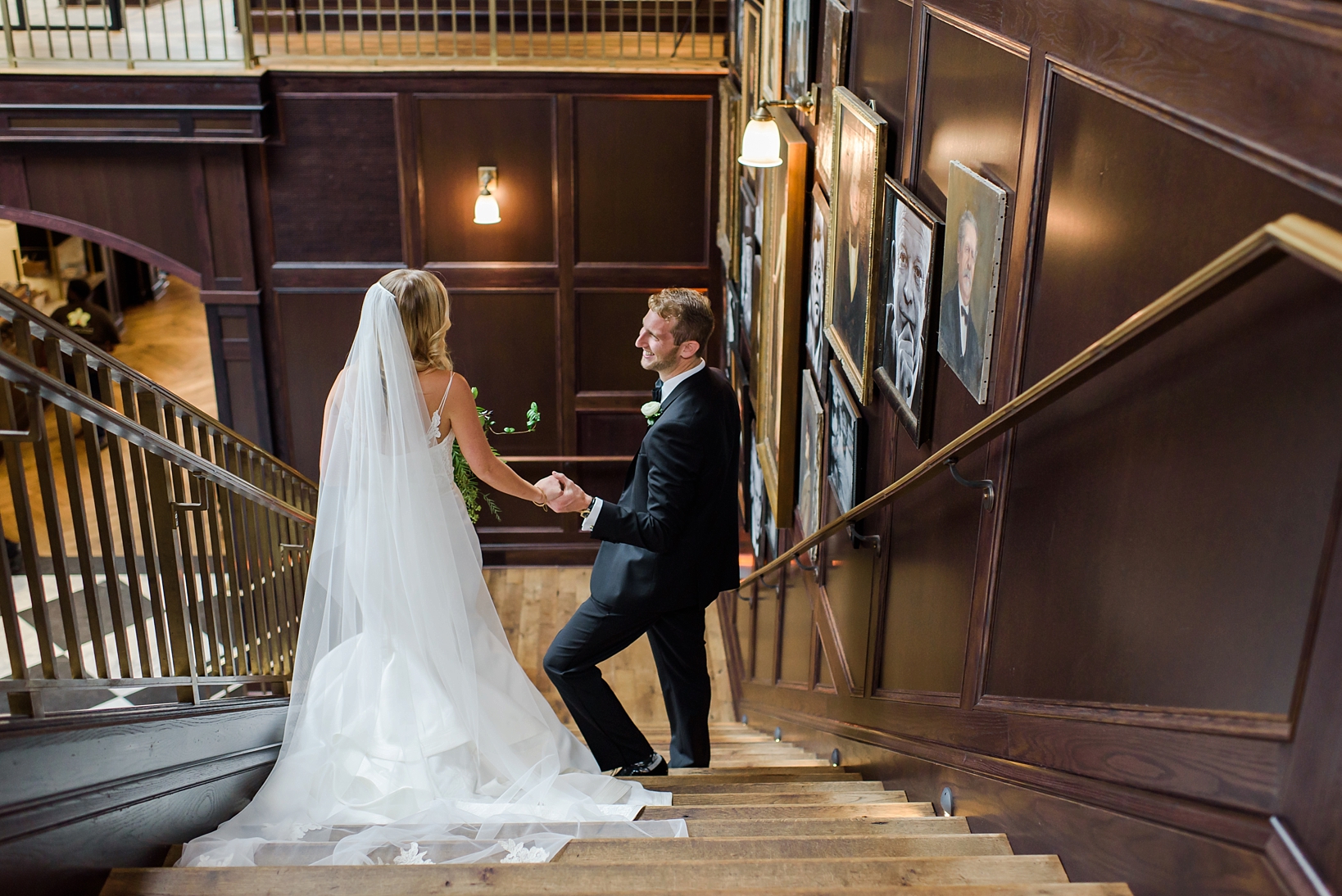 Groom helping his Bride down a flight of stairs surrounded by portraits of historical figures on the way to their Oxford Exchange wedding ceremony