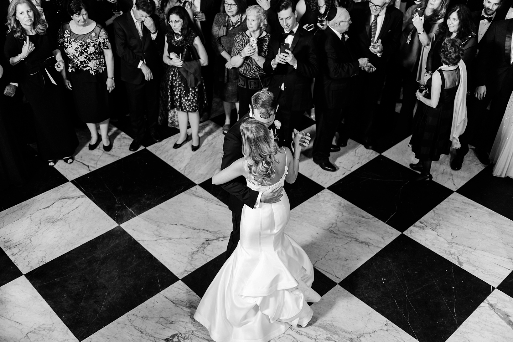 Bride and Groom share their first dance on a decorative checkered marble floor surrounded by their guests