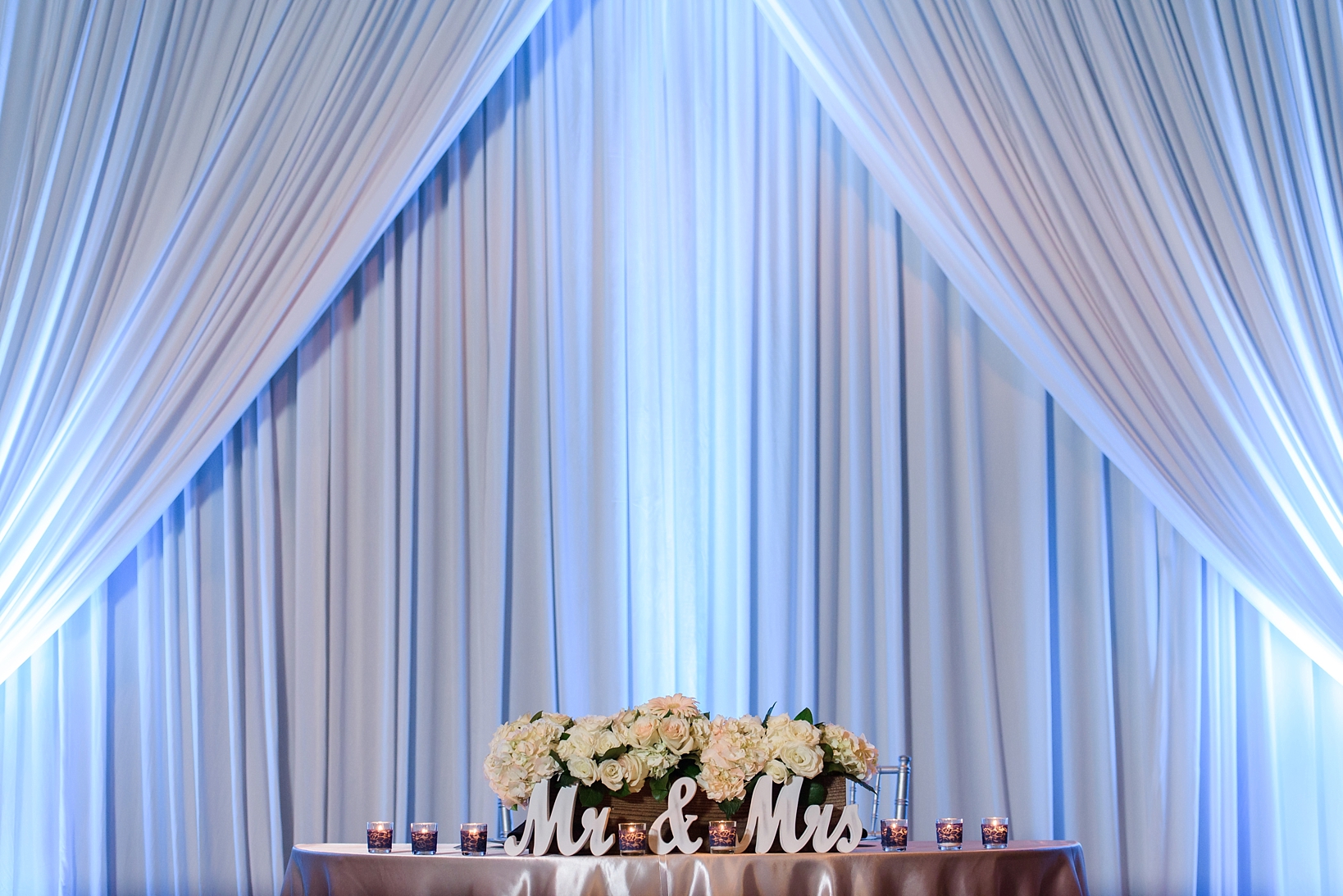 Sweetheart table peeks over the edge of the photo with blue up-lights adding personality to the drapery