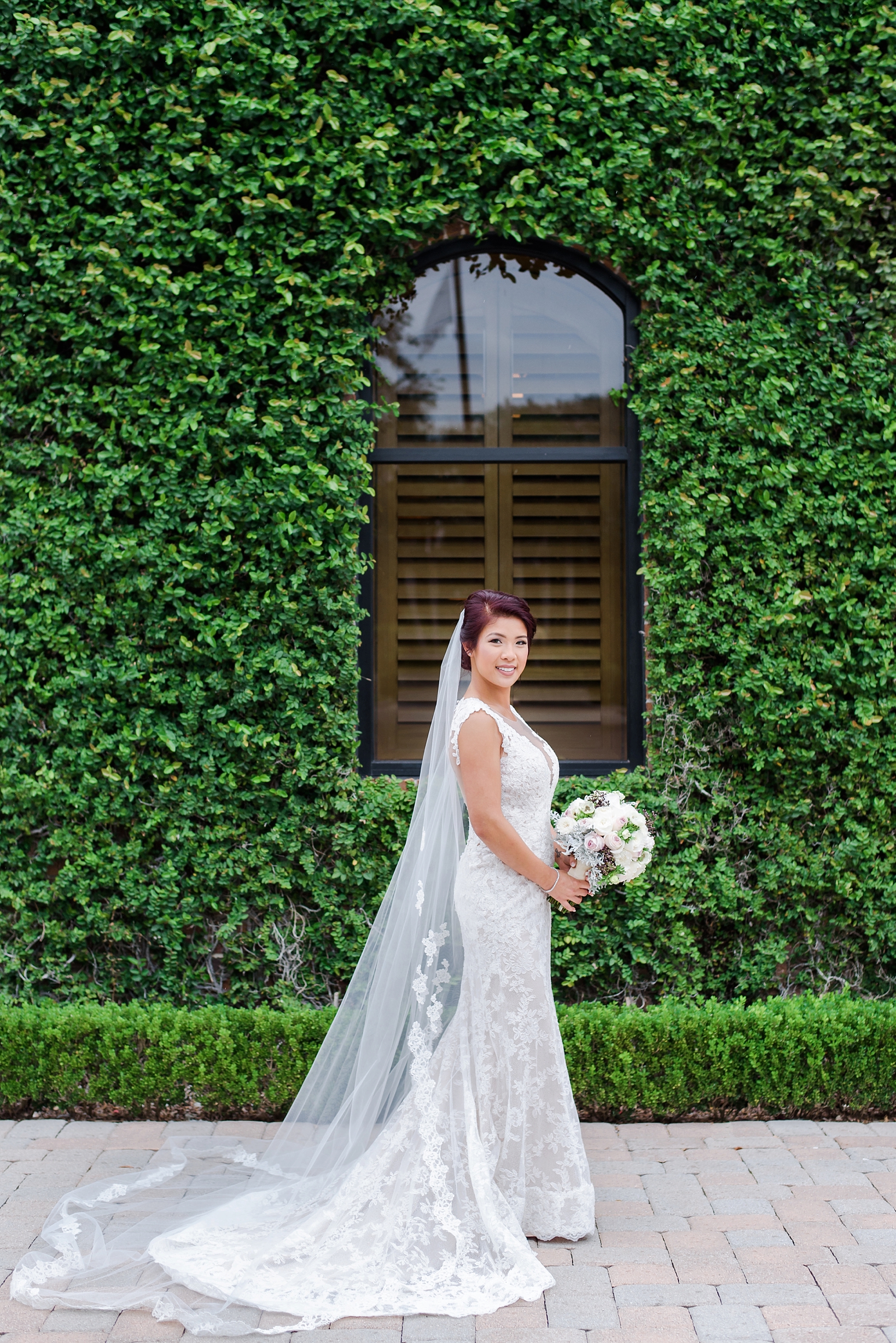 Bride with her cathedral veil and bouquet standing in front of a vine covered brick wall
