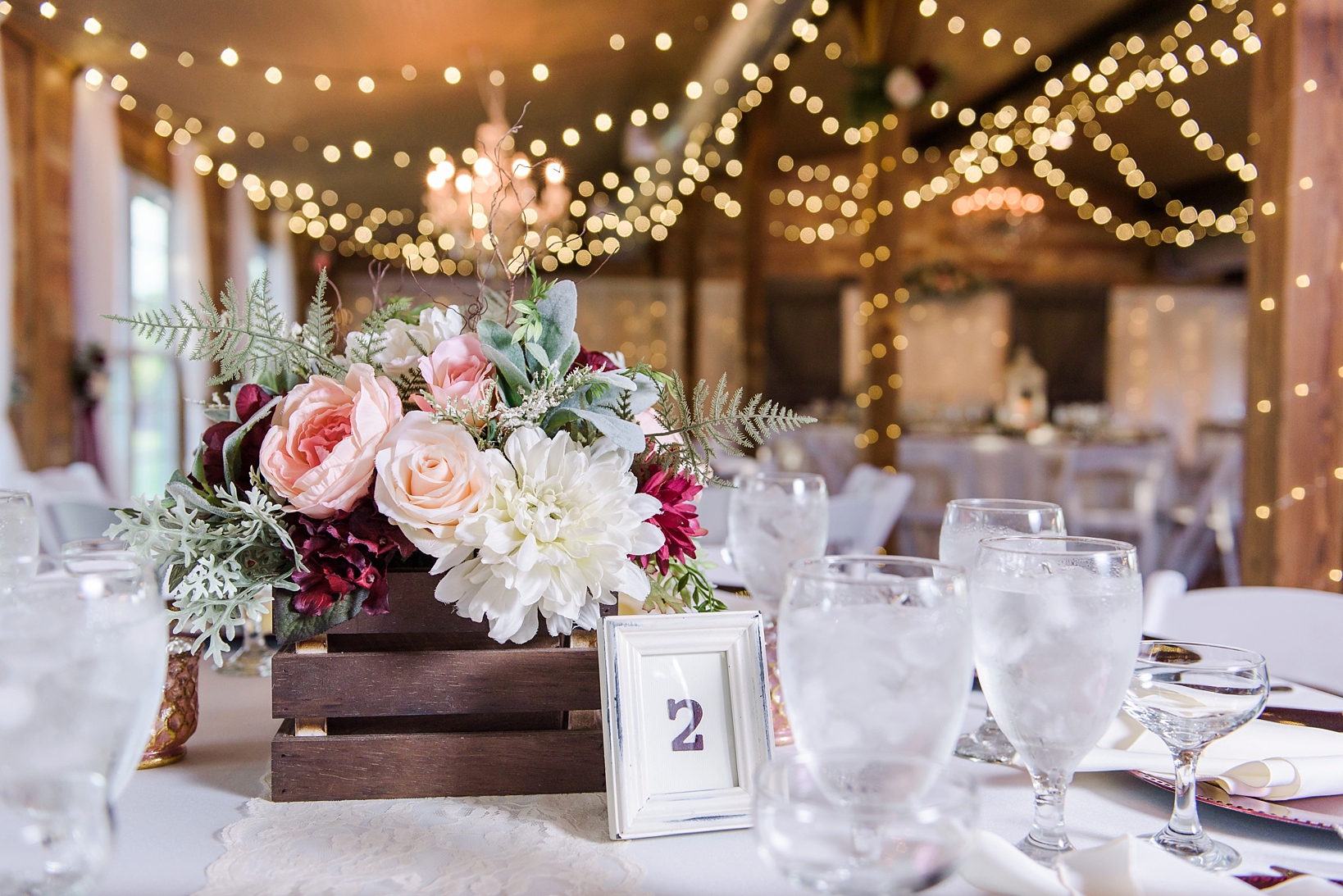 Floral centerpieces in small wooden crates surrounded by string lights in a barn
