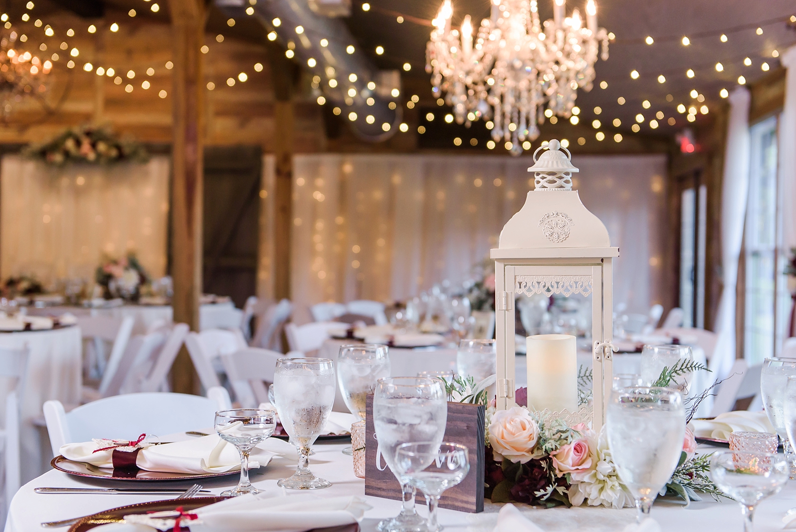 Table centerpieces of flowers and lanterns surrounded by string lights in a rustic barn setting