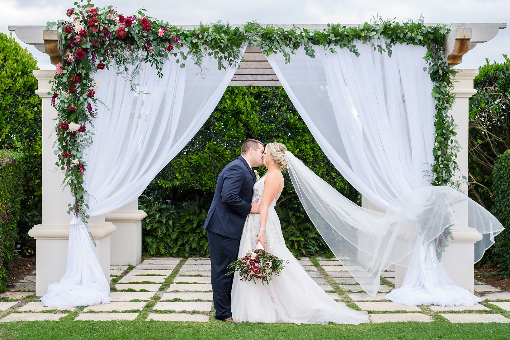 Bride and Groom kiss under their wedding arch decorated in florals as the wind blows her veil