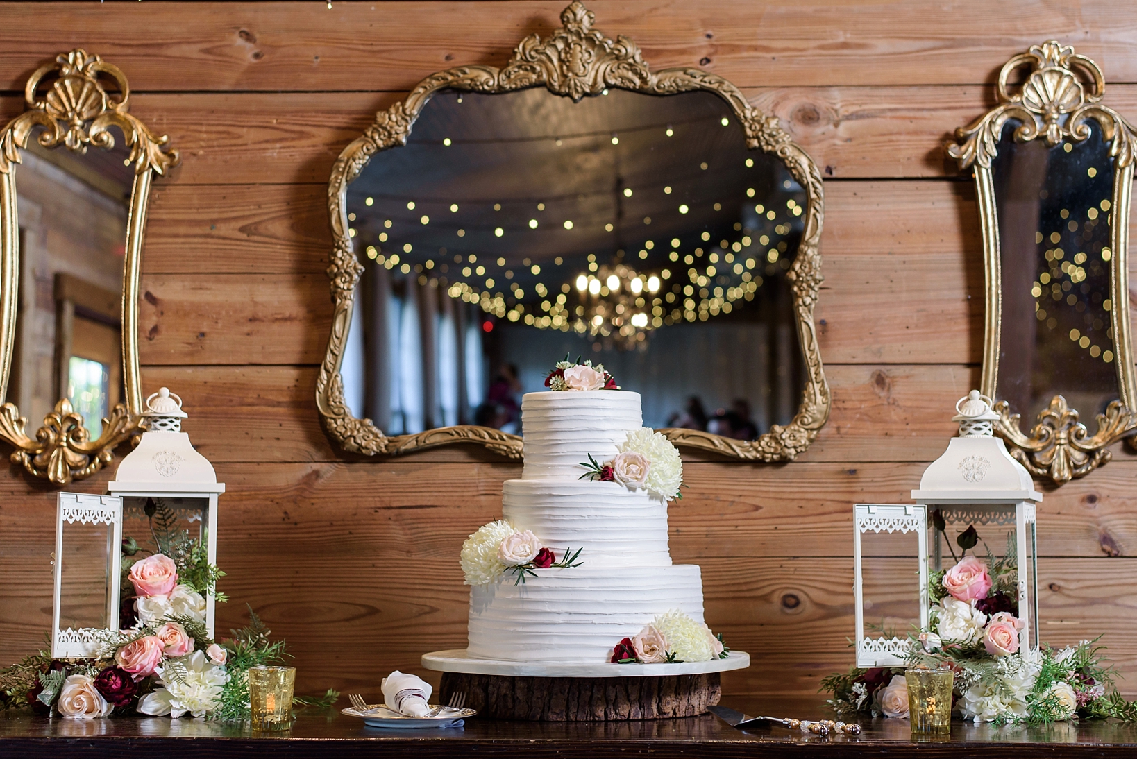 The wedding cake surrounded by reflecting mirrors and string lights with pops of floral accents