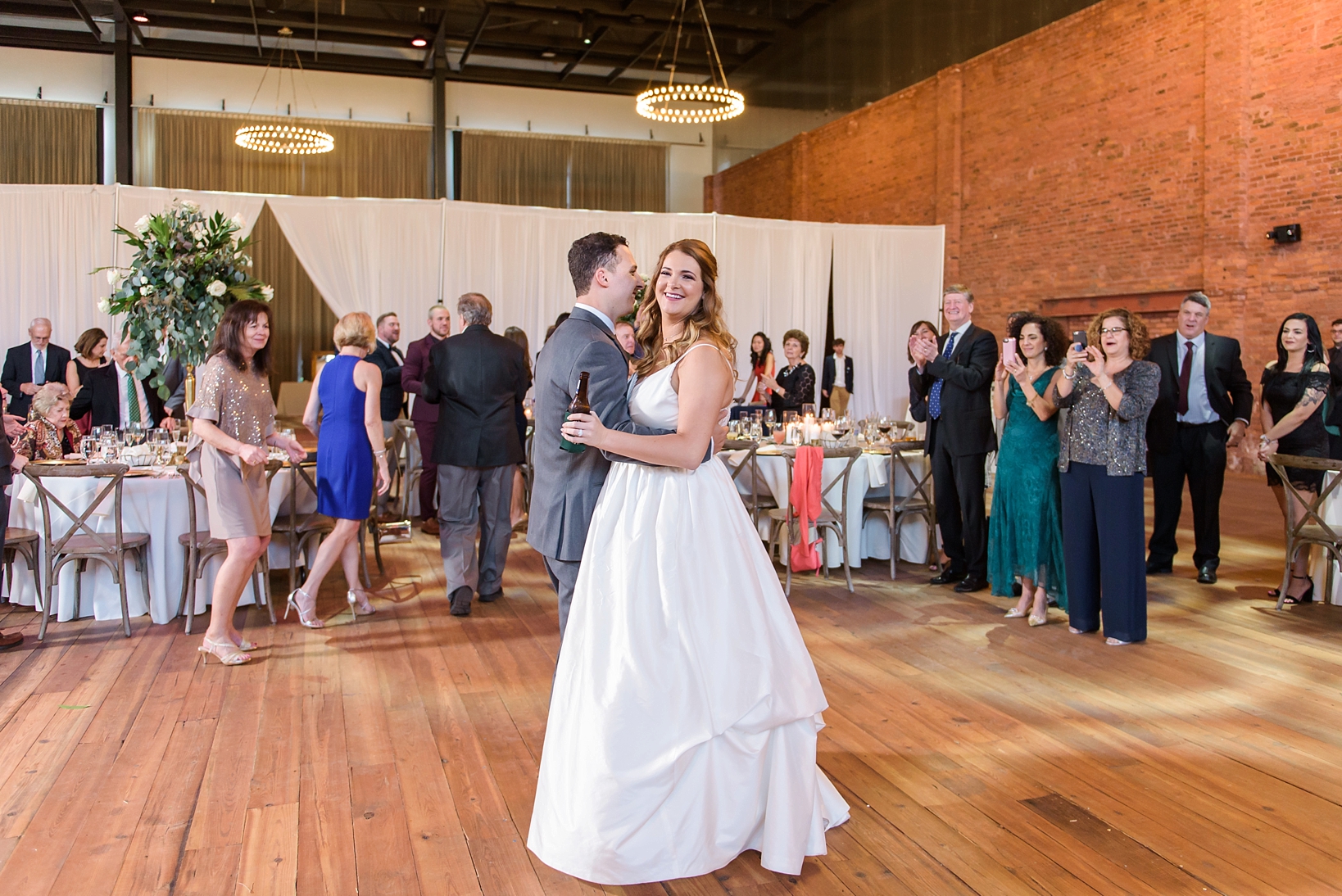 Bride and groom share their first dance while holding beer as family look on
