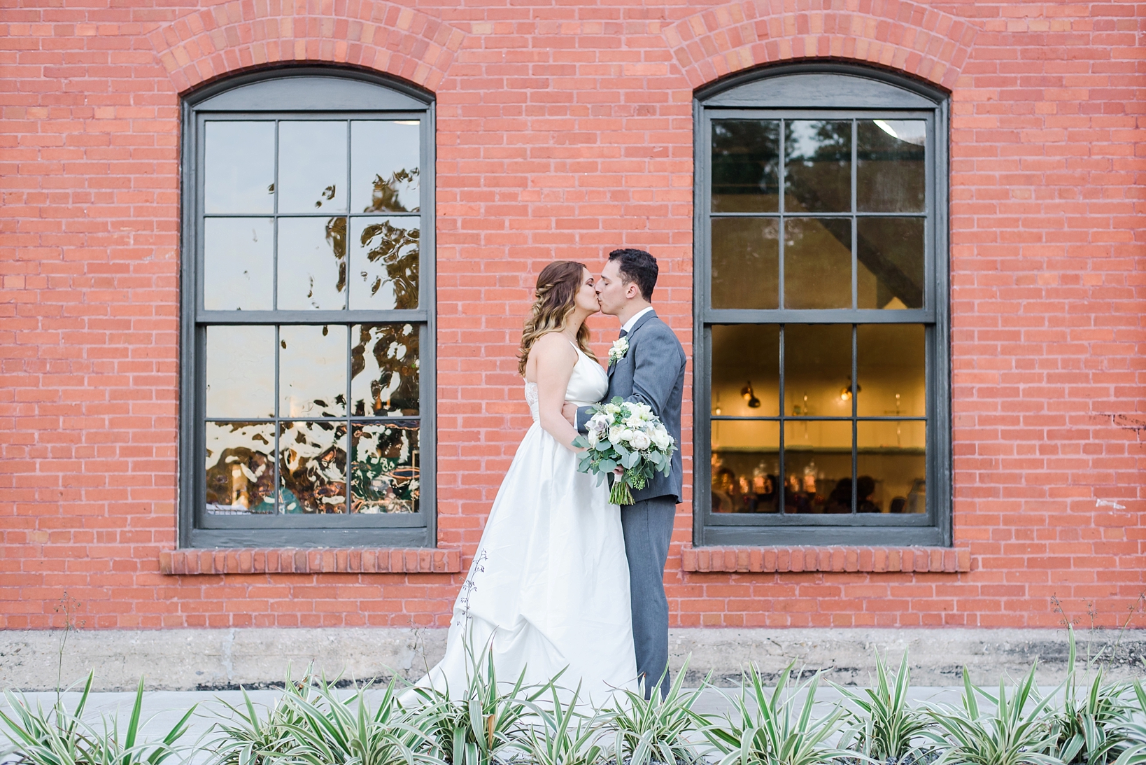 Bride and Groom share a kiss against a brick wall and industrial windows.