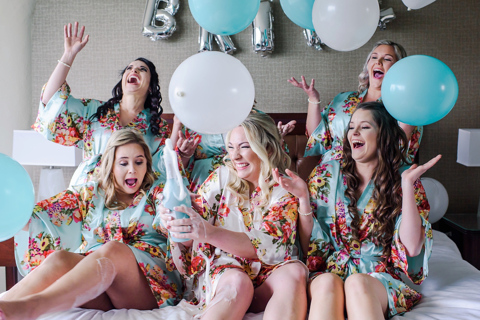 The bride and her bridesmaids popping champagne on the bed surrounded by balloons