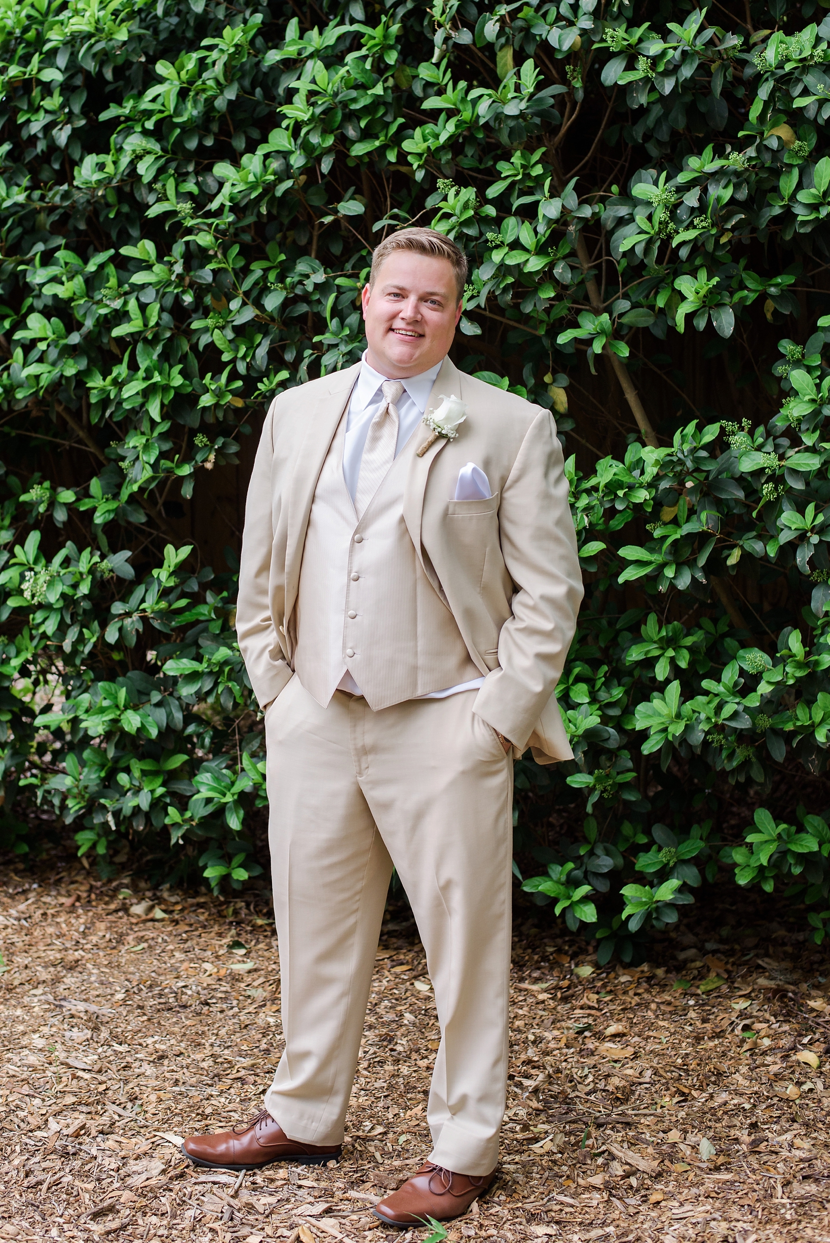The groom in a cream suit standing in front of a greenery covered wall