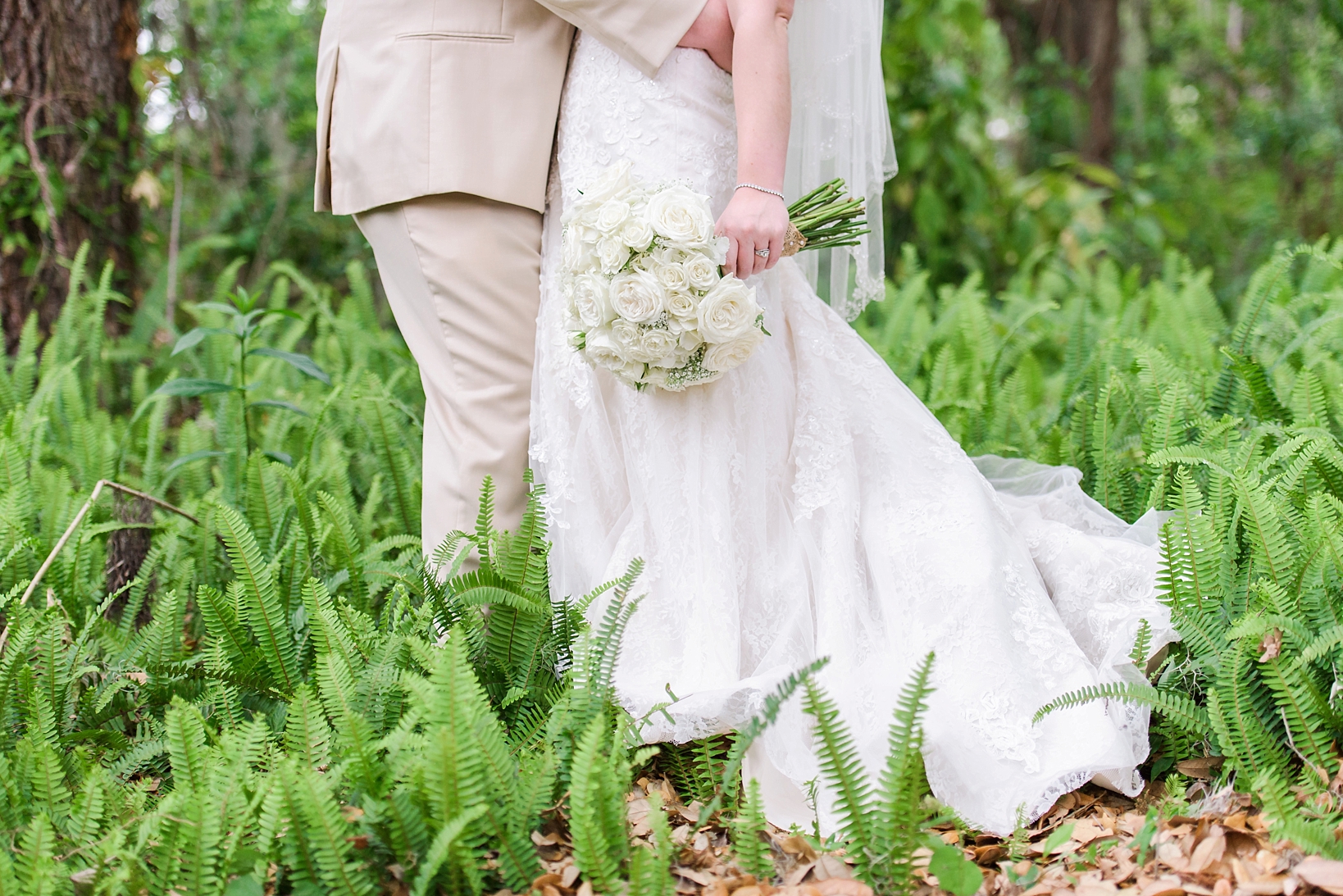 White roses wedding bouquet and groom in a cream suit surrounded by ferns