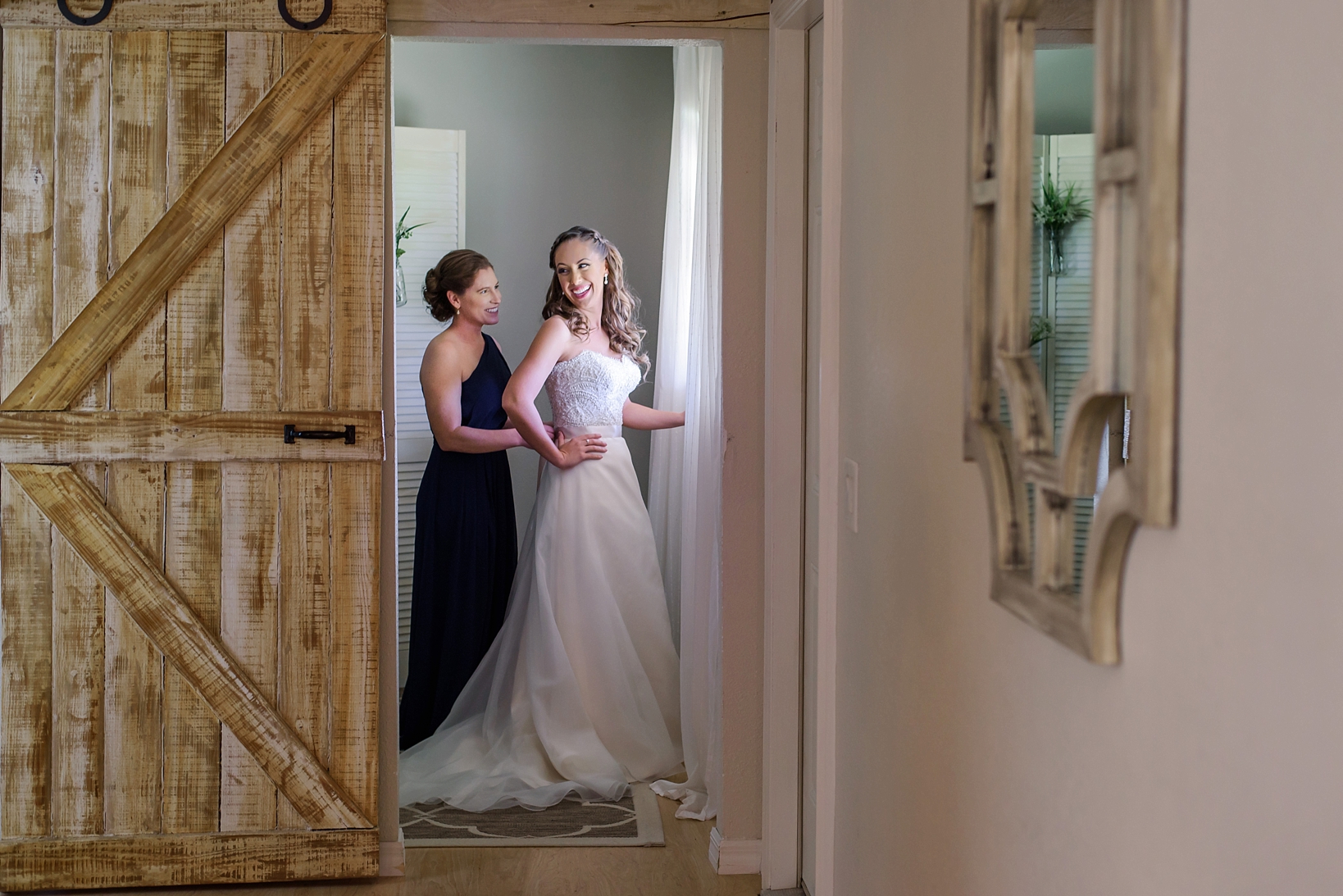 Bride being zipped into her dress by a bridesmaid inside a barn door