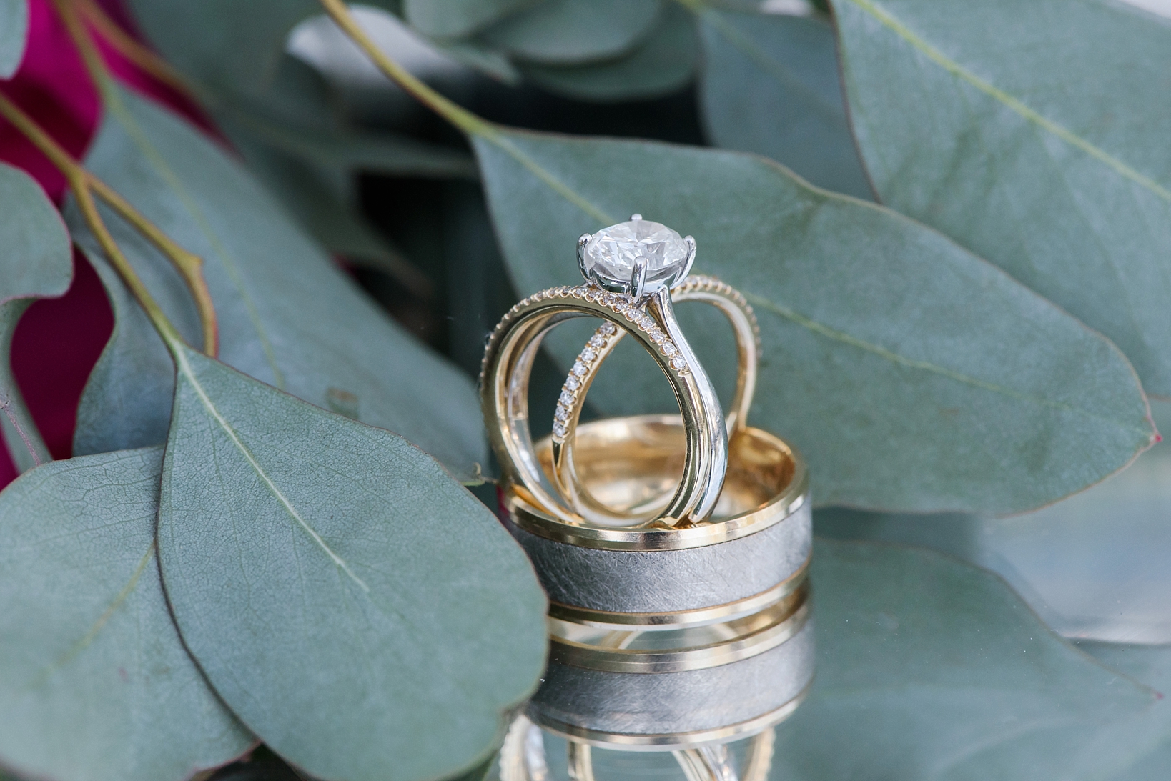 The wedding rings on a mirrored tabletop with accents of eucalyptus leaves by Sarah & Ben Photography