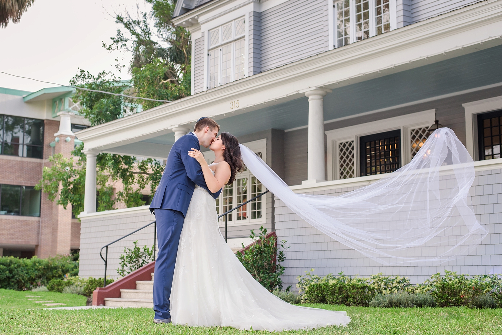 The bride and groom kiss on the front lawn as the wind sweeps the veil into the air