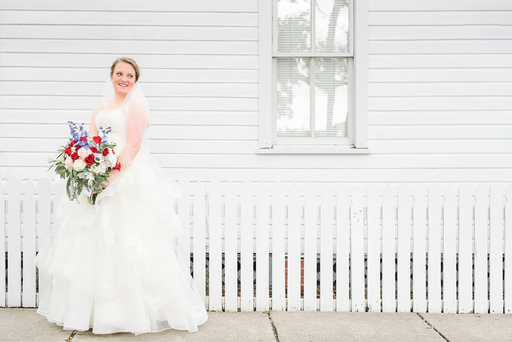 Asymmetrical portrait of a bride holding her red white and blue floral bouquet in front of a white picket fence