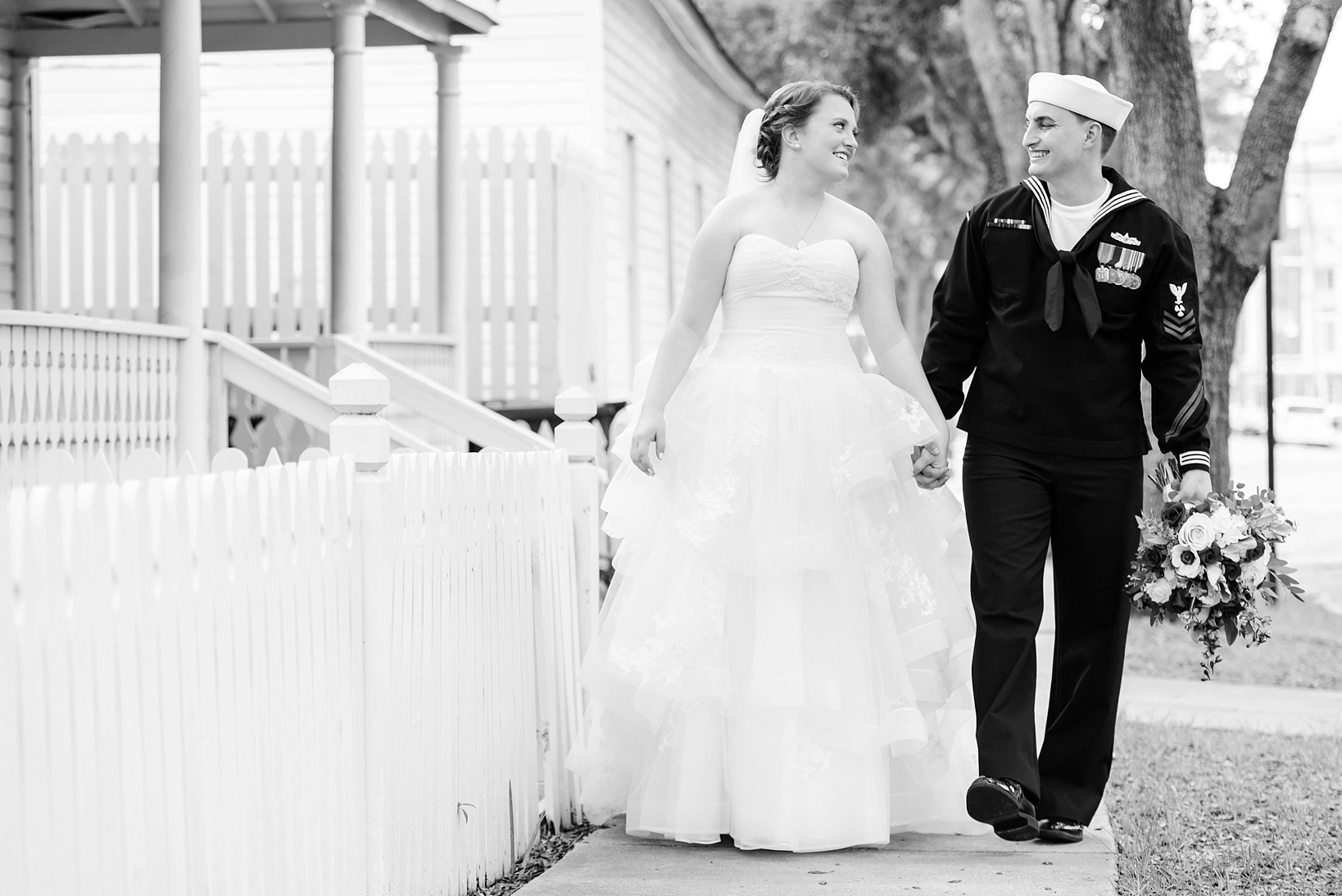 Classic black and white image of a bride and groom walking along a sidewalk next to a picket fence