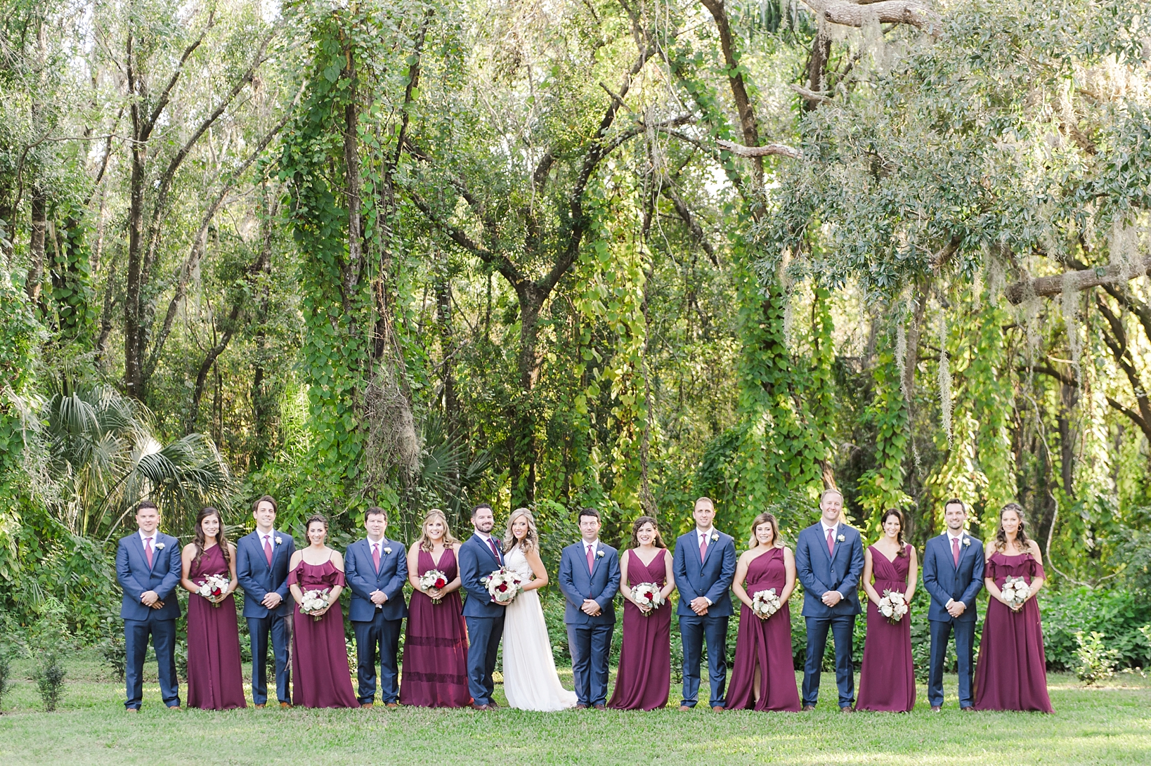 The entire wedding party wearing blue and merlot colors against a greenery-filled back ground