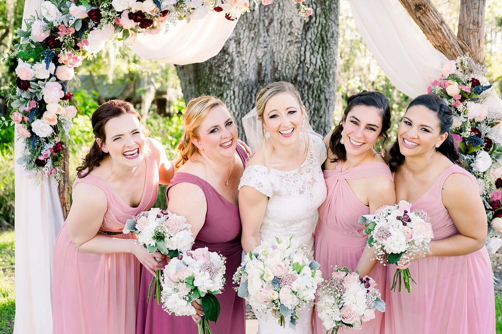 Bride and her Bridesmaids pose under the wedding arch while holding their florals