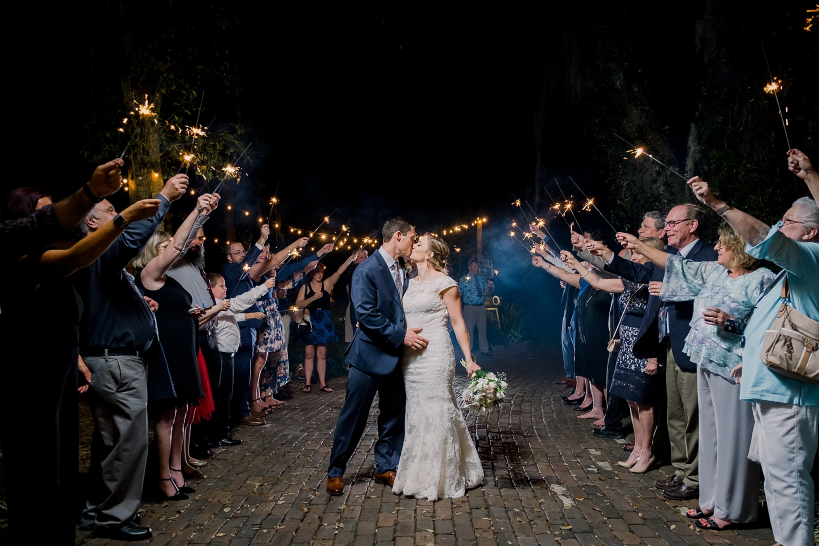 Kissing couple under a sky of sparklers held by their friends and family on the cobblestone street of Cross Creek Ranch