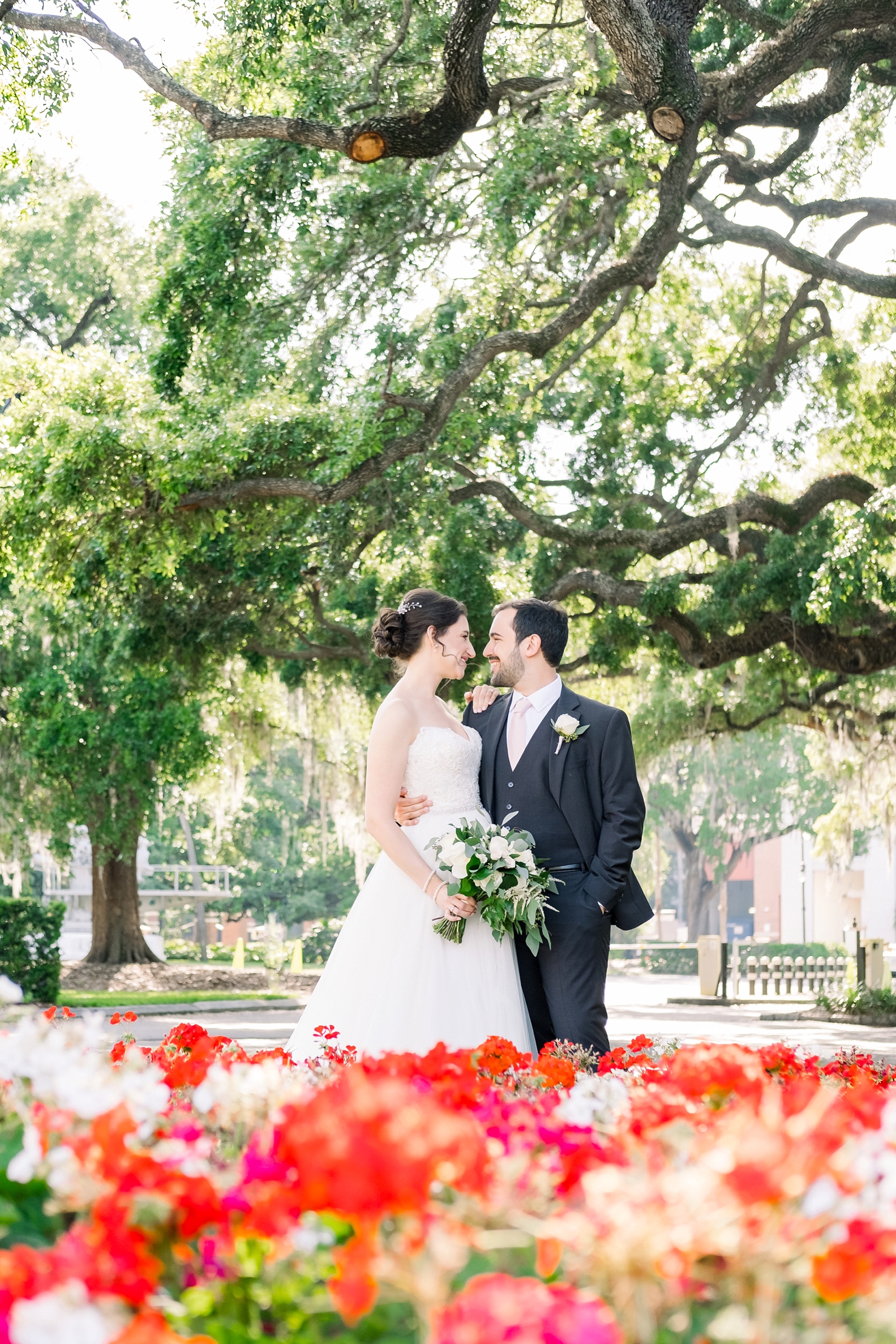 Bride and Groom under an oak tree above a bright orange flower bed in full bloom