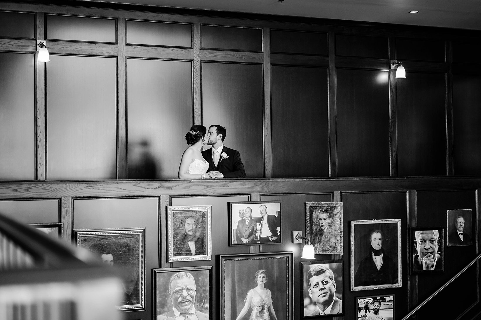 Black and White photo of the Bride and Groom kissing above portraits hanging on a wall below them
