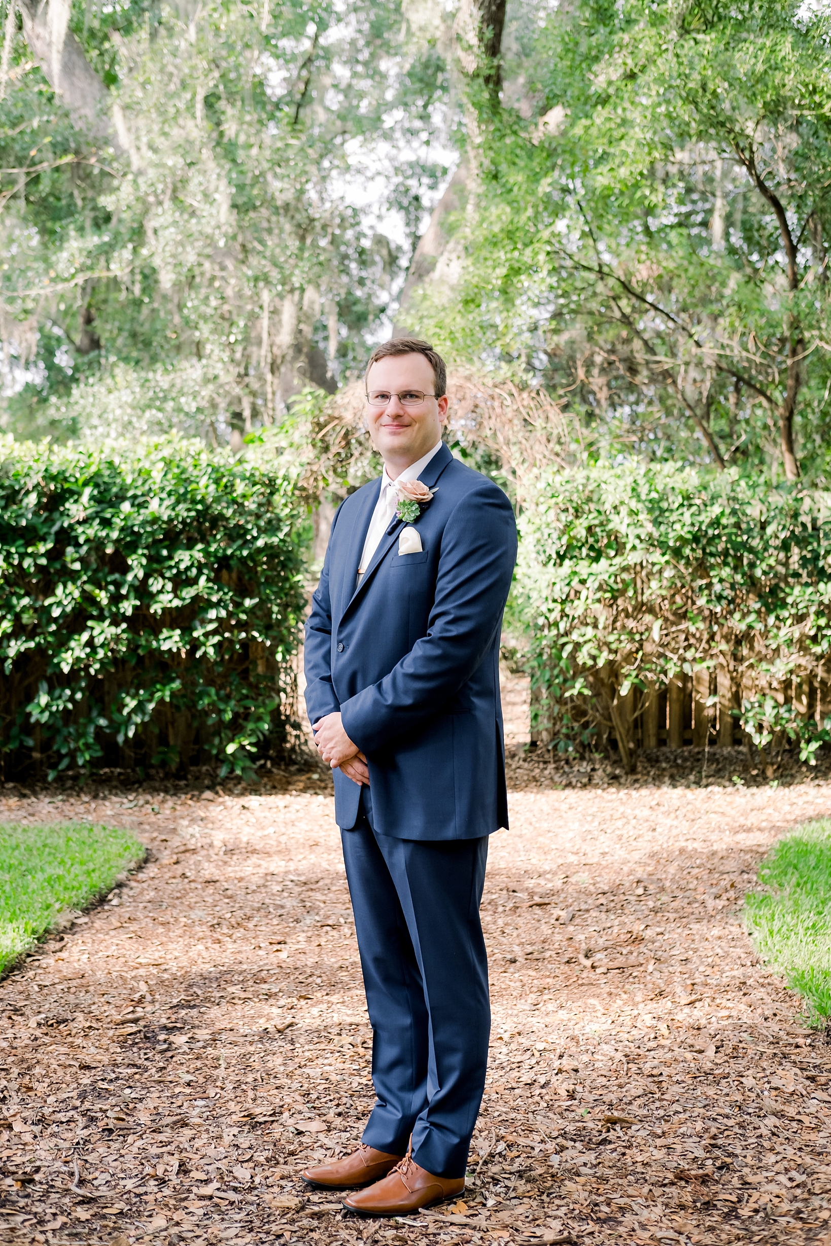 Groom stands for a formal portrait in his navy suit under the ancient oak trees of Cross Creek ranch