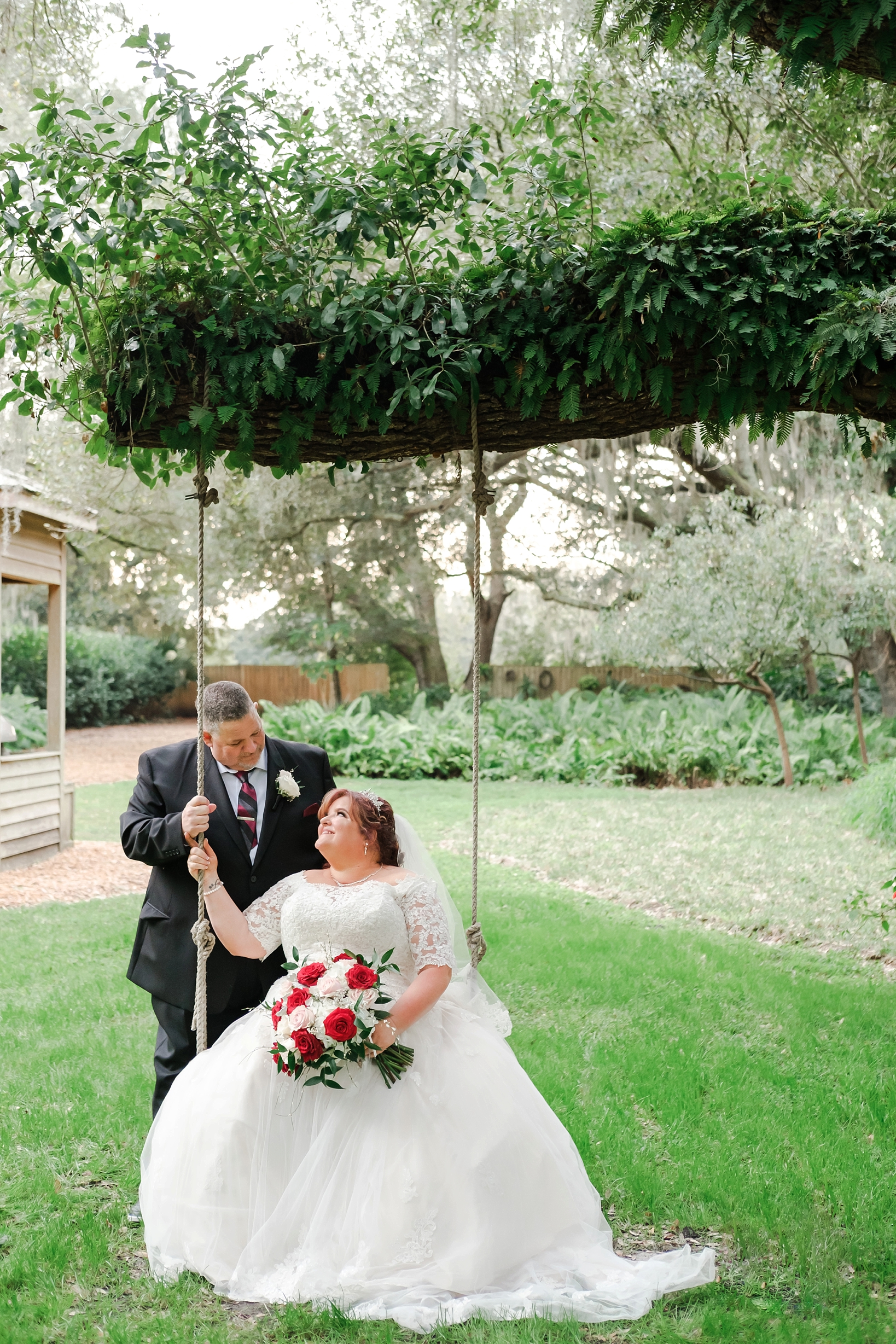 Bride sits on a swing as her Groom stands behind her by Sarah and Ben Photography