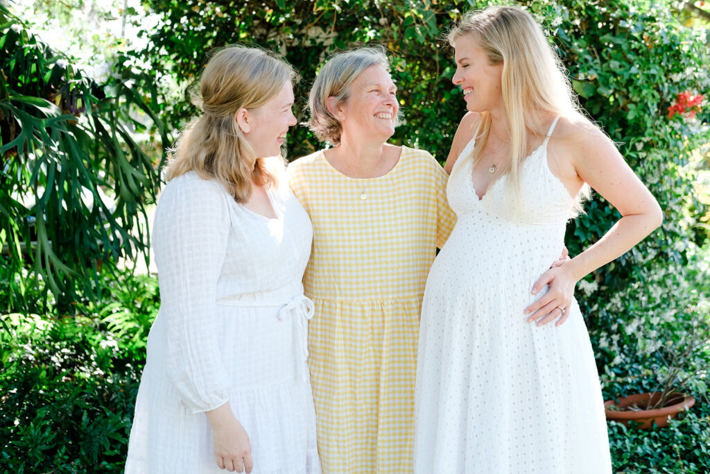 A Mother smiles while holding her two adult Daughters against a floral backdrop
